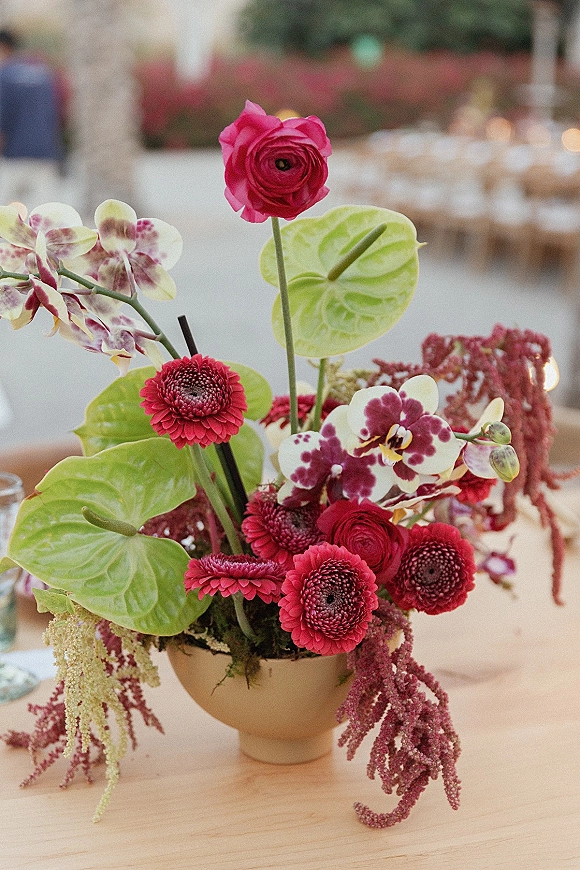 Wedding centerpiece with modern floral centerpiece styling, featuring red ranunculus, gerbera daisies, orchids, and anthurium in a ceramic bowl under bistro lights