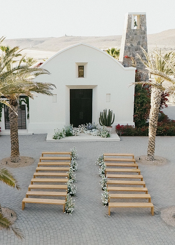 Ceremony setup with wood benches and white and green aisle florals leading to a white chapel facade with palm trees and a stone bell tower backdrop