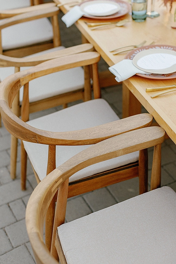 Reception tablescape on a wood wedding table with terracotta chargers, white napkins, gold flatware, water glasses, and cushioned chairs on a stone patio
