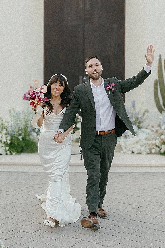 Recessional moment as bride and groom recessional hand in hand, groom waving, bride with bouquet at double doors with cacti backdrop