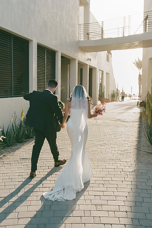 Couple portrait of bride and groom holding hands, walking away in golden light, her long veil trailing beside a modern building.
