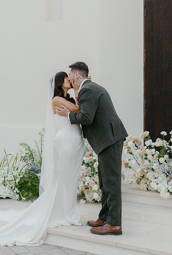 Wedding kiss portrait of bride and groom kissing, her veil flowing as she holds his face, by a white wall and dark wood door