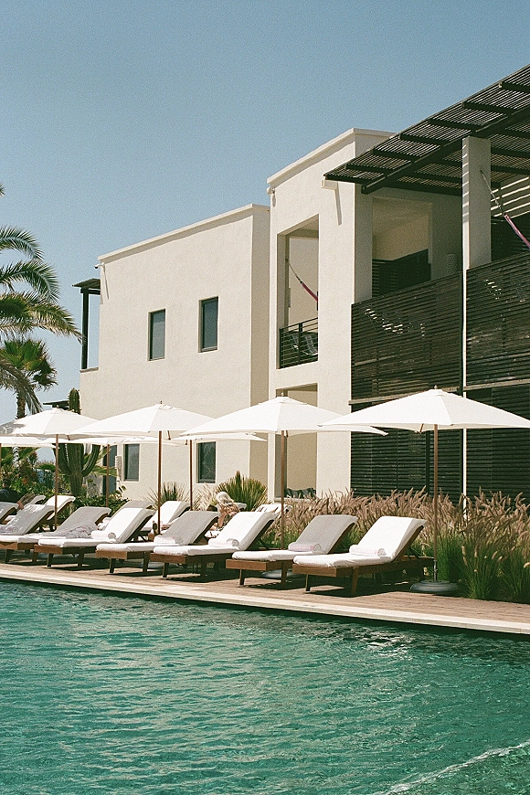 Resort poolside with poolside lounge chairs beside clear water and white patio umbrellas, set against a modern white building and palms