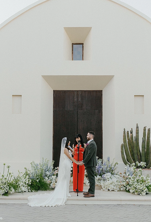Wedding vows during an outdoor wedding ceremony as bride and groom hold hands before a white stucco chapel with cactus accents
