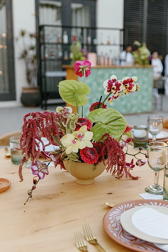Reception tablescape with wedding table centerpiece of orchids, anthurium leaves, and amaranthus in a ceramic bowl on an outdoor patio bar backdrop
