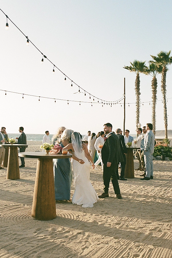 Wedding cocktail hour on the beach with guests mingling by high-top tables under string lights, floral centerpieces, ocean and palms behind