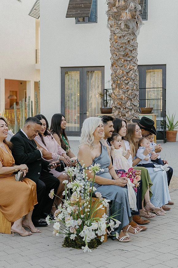 Wedding guests seated in ceremony guest seating, smiling in pastel dresses and a cowboy hat amid crate florals in a sunny courtyard