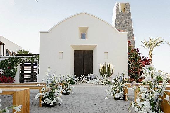 Ceremony setup with outdoor ceremony setup aisle florals lining a courtyard leading to a white chapel facade and stone bell tower backdrop