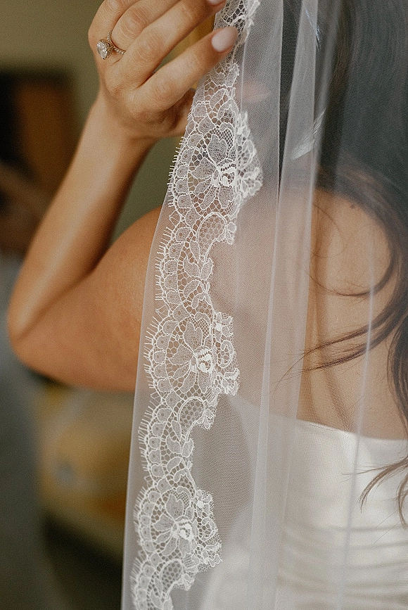 Bridal veil detail with a lace edge veil draped in soft light, bride’s manicured hand showing an engagement ring over tulle