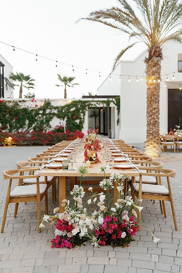 Reception tablescape with an outdoor wedding reception table on a wood banquet table, candles and florals under string lights in a palm courtyard