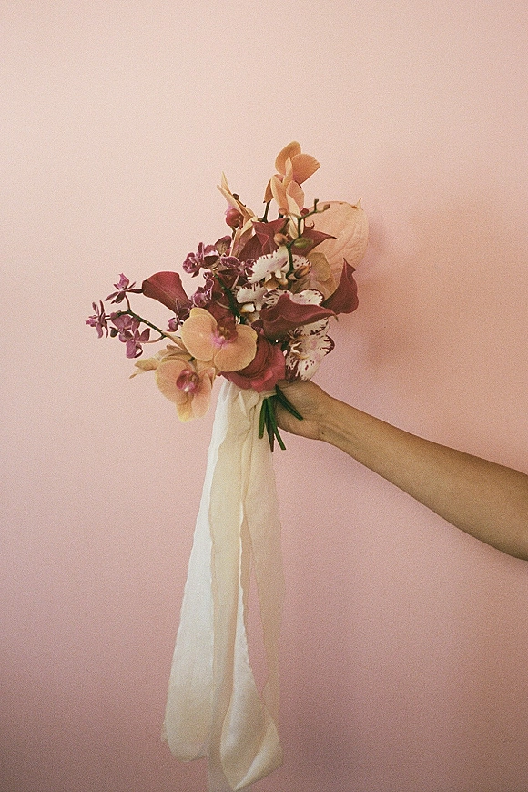 Bridal bouquet with orchid bridal bouquet blooms, calla lilies, greenery, and long trailing ribbons held in hand against a pink wall backdrop