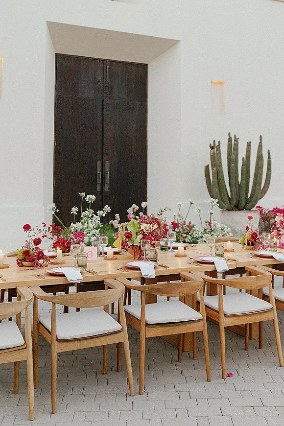 Reception tablescape on a wood banquet table wedding setup with white napkins, candles, colorful florals, and cactus courtyard backdrop.