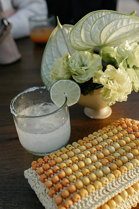 Wedding cocktail with salt rim and lime garnish over ice beside anthurium and carnation centerpiece on a wood table with blurred guests
