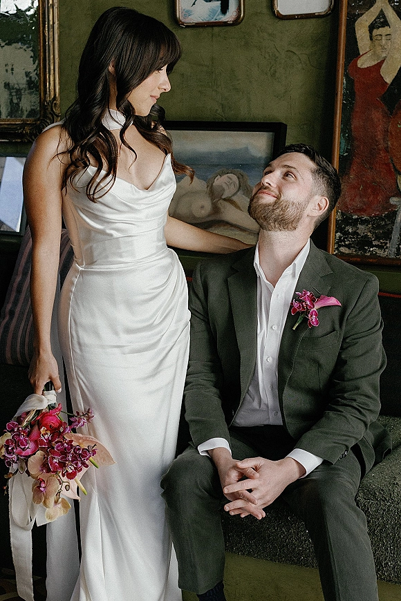 Couple portrait of bride in satin dress holding an orchid bouquet beside her seated groom, against a green gallery wall with framed art