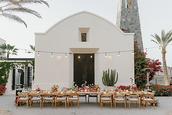 Reception tablescape with a long banquet table wedding setup, floral centerpieces, candles, and string lights in a chapel courtyard backdrop