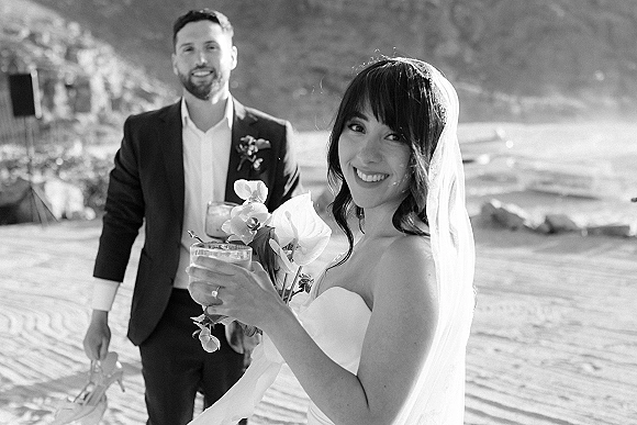 Couple portrait at a beach wedding portrait, bride smiling back in veil and strapless dress as groom holds cocktails by rocky cliffs and waves