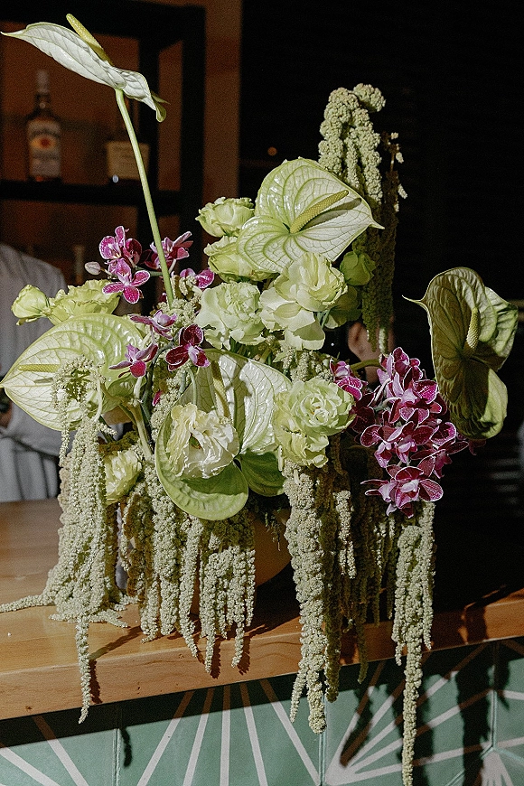 Wedding floral centerpiece with anthurium and orchids, white roses, and hanging amaranthus in a vase on a dimly lit reception bar counter