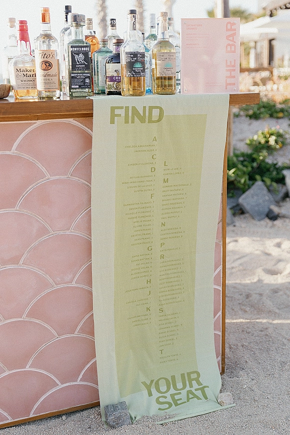 Wedding seating chart alphabetical fabric banner hanging over a wooden bar with liquor bottles and signature drinks sign in sunlit greenery