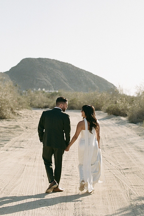 Couple portrait of bride and groom holding hands, walking away on a desert road with mountains behind, veil flowing in the wind