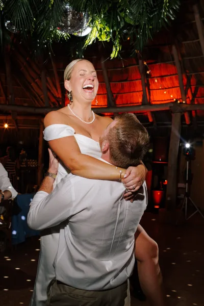 Reception dance moment as groom lifts a laughing bride in an off the shoulder dress beneath disco ball and string lights on a crowded dance floor