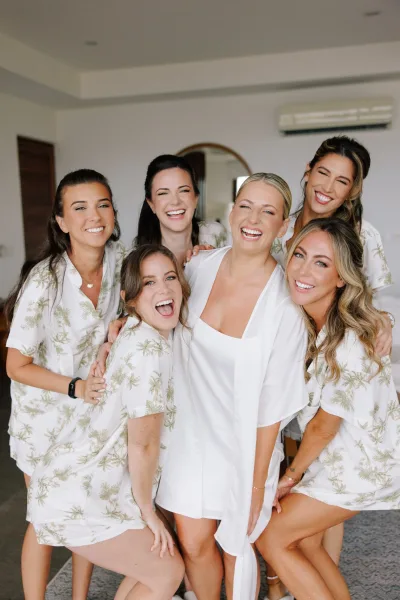 Bridesmaids getting ready in bridal party pajamas, laughing beside the bride in a white satin robe in a hotel room with mirror backdrop