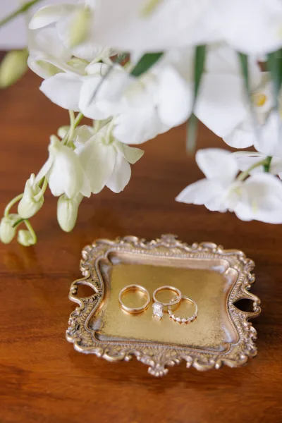 Wedding rings arranged on an ornate gold tray with a white orchid accent, set on a rustic wood table in close-up detail