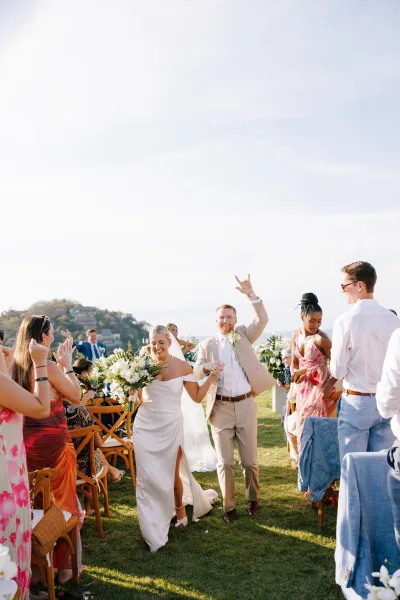 Ceremony recessional as bride and groom walk up aisle, bouquet and veil flowing, guests cheering on a sunny hillside with ocean view