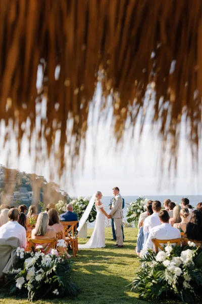 Wedding ceremony with bride and groom holding hands under a white floral arch, ocean view beyond guests on a grassy cliffside lawn