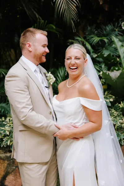 Couple portrait of bride and groom laughing while holding hands, her long veil and pearl jewelry beside his beige suit in tropical greenery