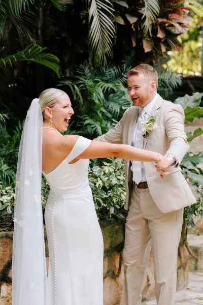 First look moment as bride and groom laugh holding hands, her long veil flowing beside his beige suit amid tropical palm greenery
