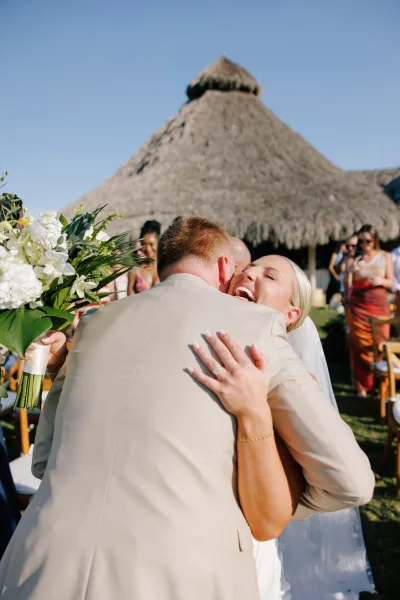 Ceremony hug as bride and groom embrace, bride laughing with bouquet and veil, guests on lawn under thatched roof, blue sky
