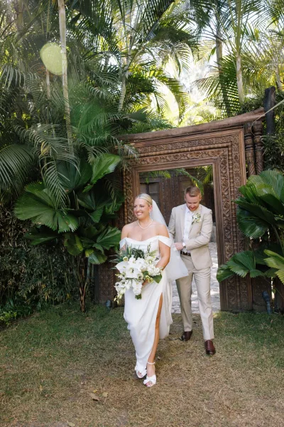First look moment as groom sees bride in off-shoulder dress and veil holding white bouquet, by ornate carved wooden doors in sunlit tropical garden