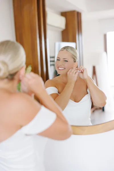 Bridal getting ready bride putting on earrings in a mirror, wearing an off-the-shoulder wedding dress in a bright, wood-paneled room