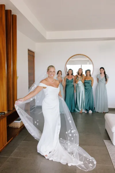 Bride portrait in an off the shoulder wedding dress with a long veil and pearl necklace, standing in a modern bridal suite with white walls and mirror