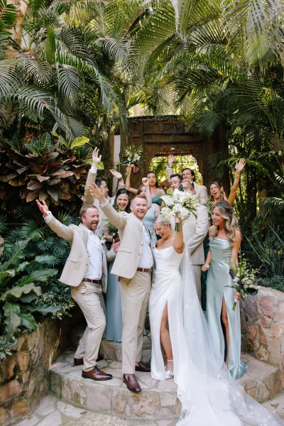 Wedding party portrait with the bride and groom with wedding party cheering on stone steps, bouquet raised, amid tropical palm greenery and carved doorway