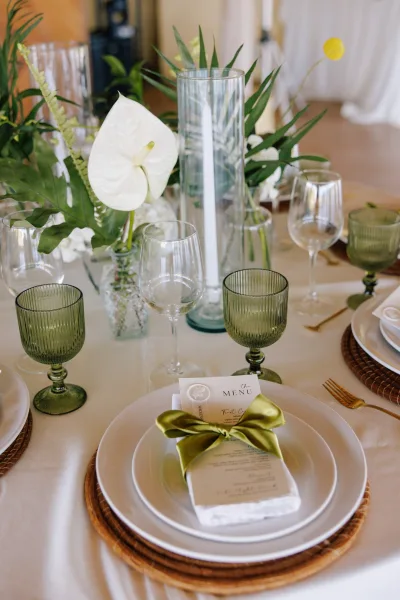 Reception tablescape with wedding place setting on rattan charger, green goblets, gold flatware, ribbon menu, and taper candles under soft drapery