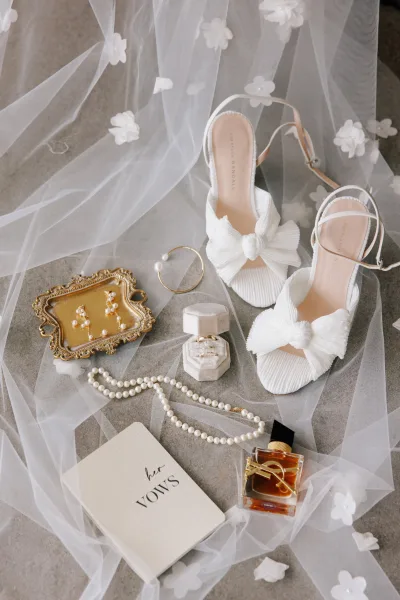 Bridal accessories flatlay with white bow heels, tulle veil with floral appliques, pearls, ring box, perfume and vow book on gray fabric background