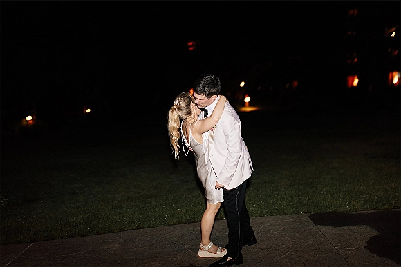 Wedding kiss as bride in a satin mini dress embraces groom in a white tuxedo jacket on a candlelit walkway under the night sky
