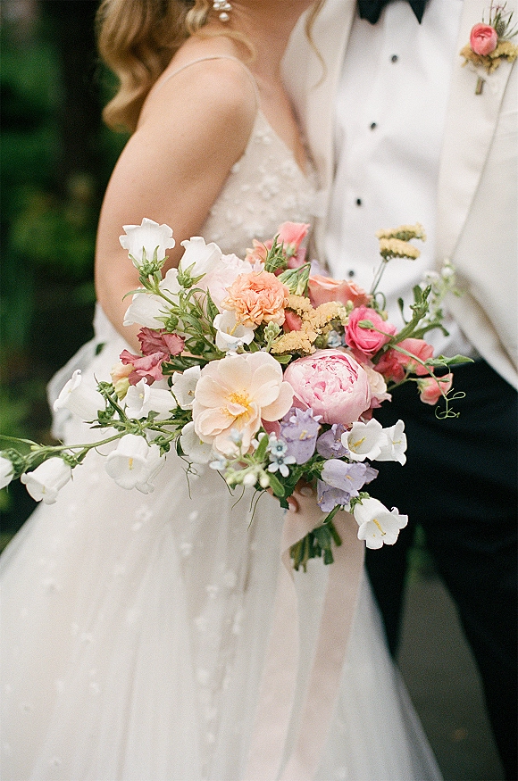 Bridal bouquet with pink peony bouquet blooms and ranunculus, held by bride in gown beside groom in white tuxedo, garden greenery behind