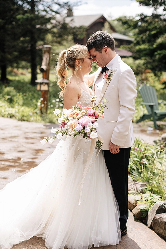 Couple portrait of bride and groom forehead touch, bride holding pink-and-white bouquet on a stone path amid garden greenery and trees