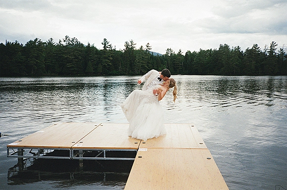 Wedding kiss portrait of bride in a strapless gown and groom in a white tuxedo dipping on a dock by a mountain lake under cloudy sky