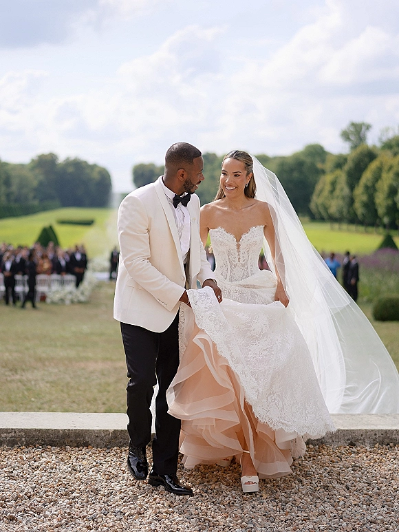 Wedding couple portrait of bride and groom walking down an outdoor aisle, bride laughing in long veil and lace gown as guests blur behind