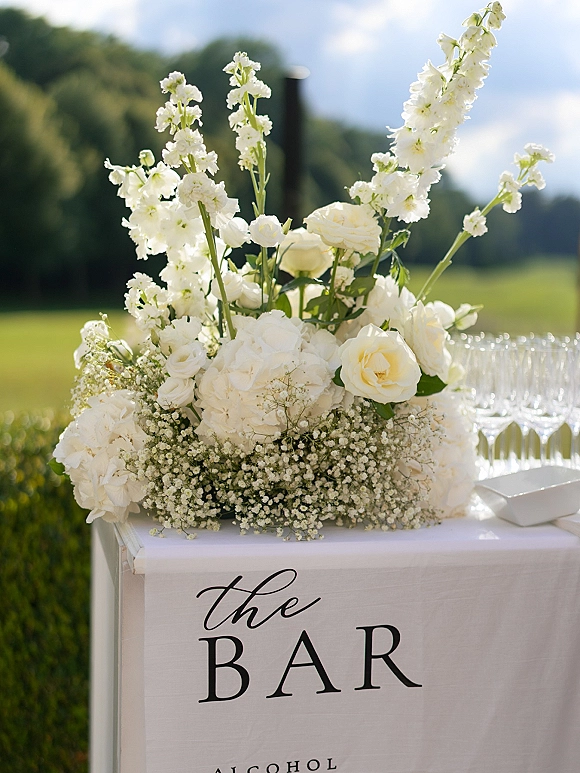 Wedding bar sign with elegant calligraphy beside champagne glasses and a white rose and hydrangea arrangement on a lawn setting