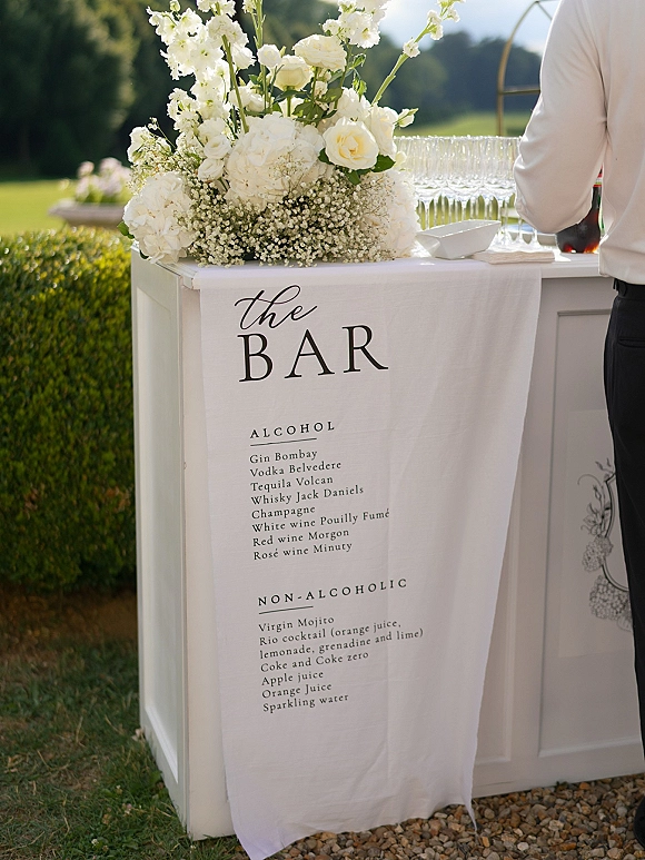 Wedding bar sign wedding bar menu on a linen banner with calligraphy, framed by white roses and hydrangeas on an outdoor bar counter