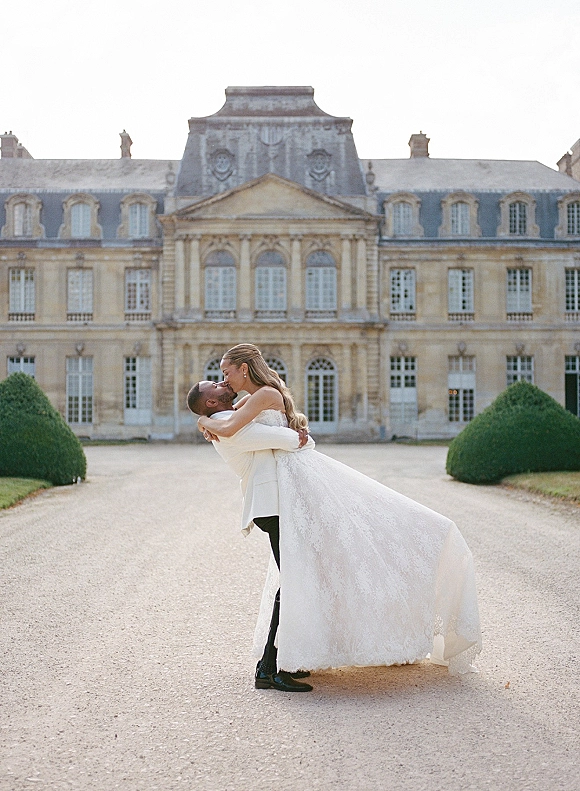 Wedding kiss portrait of groom lifting the bride in a dip, her strapless lace gown train flowing in a gravel estate courtyard