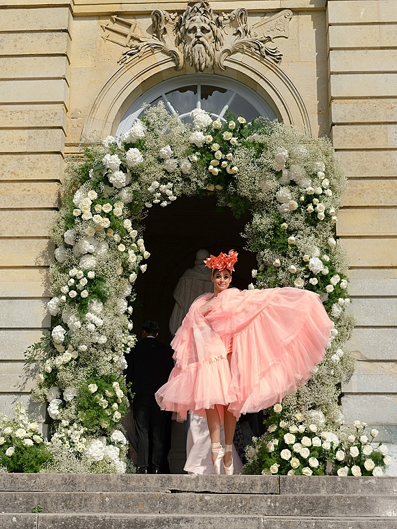 Bridal fashion portrait of a bride in a pink tulle bridal gown with floral headpiece, standing on stone steps beneath a white rose arch