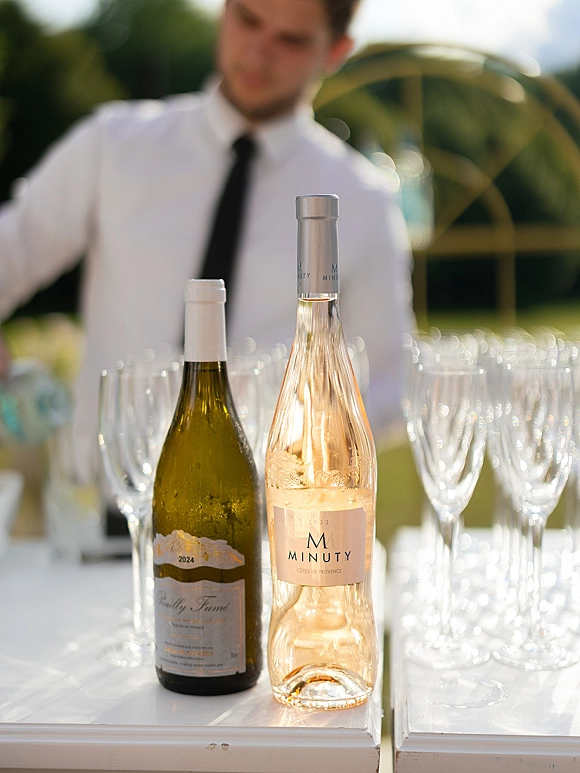 Wedding bar setup with wine bottles and champagne flutes lined on a bar table, with bartender on an outdoor lawn beneath a circular metal arch