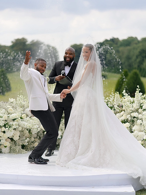 Ceremony moment as bride and groom hold hands at an outdoor wedding ceremony, officiant with microphone, white roses and fountain behind