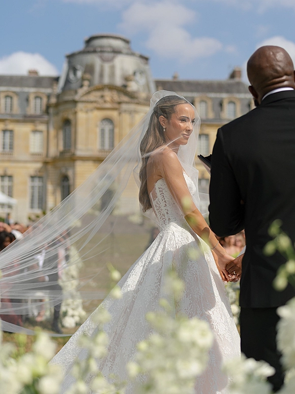 Ceremony moment as bride and groom hold hands, her long veil flowing in the wind along a floral aisle in a historic courtyard