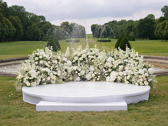Wedding ceremony backdrop with a white floral arrangement of roses, baby’s breath, and greenery on a round stage by a fountain in a garden lawn